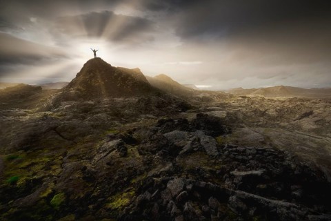 Man on a hill in a black lava field with green moss stretching hands into the dramatic sky