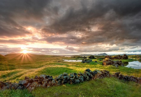 Stone wall and lava formations of volcanic rock rise from green meadows with dramatic sky and sun star in the evening light at Kalfastroend