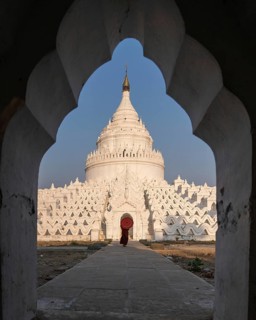 Buddhist monk stands with red umbrella in front of Hsinbyume Pagoda with frame of another pagoda