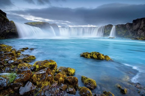 Long time exposure of a turquoise waterfall in volcanic landscape with dramatic clouds and green moss on rocks