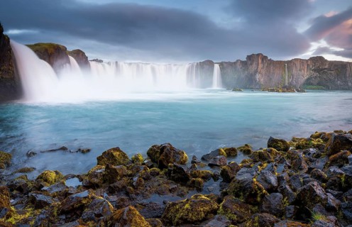 Long time exposure of a turquoise waterfall in volcanic landscape with dramatic clouds and green moss on rocks