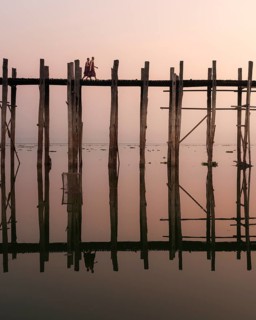 Buddhist monks walk across the U-leg bridge in red robes as the sun rises