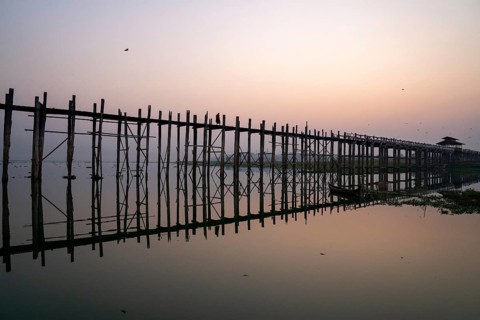 Buddhist monks walk across the U-leg bridge in red robes as the sun rises