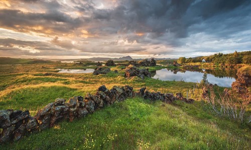Stone wall and lava formations of volcanic rock rise from green meadow with dramatic sky in the evening light at Kalfastroend