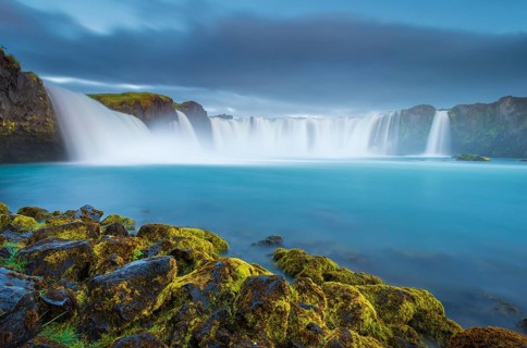 Long time exposure of a turquoise waterfall in volcanic landscape with dramatic clouds and green moss on rocks