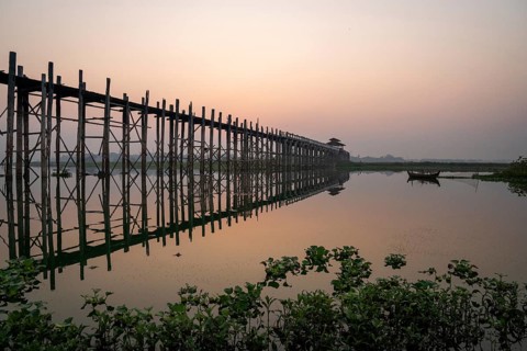 Buddhist monks walking across the U-leg bridge as the sun rises with green plants in the water