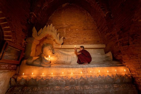 Buddhist young monk in red robe with red umbrella praying with candle in hand in a temple