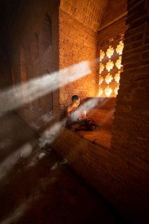 Buddhist young monk in red robe reading sitting in front of rays of light in a temple