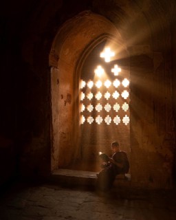 Buddhist young monk in red robe reading sitting in front of rays of light in a temple