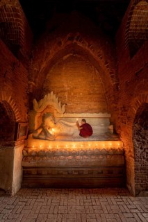 Buddhist young monk in red robe with red umbrella praying with candle in hand in a temple