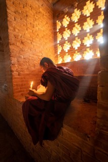 Buddhist young monk in red robe reading while sitting with a candle in front of rays of light in a temple