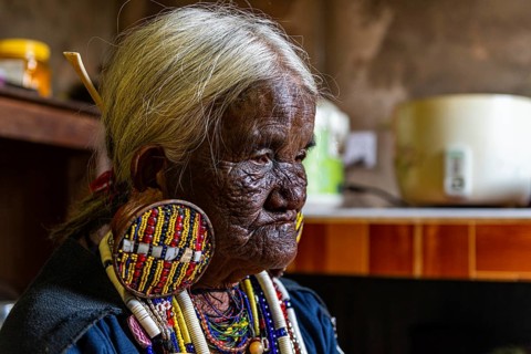 Chin Kaang hill-tribe woman with traditional facial tattoo and elaborate tribal earrings playing a flute with her nose
