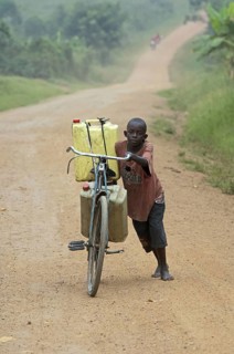 Boy pushing bicycle laden with full water containers along dusty road