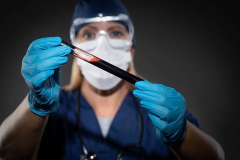 Female Lab Worker Wearing Medical Face Mask Holds Test Tube of Blood Against Dark Background