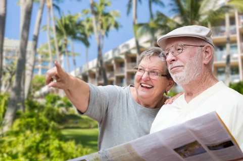 Happy senior adult couple tourists with brochure next to tropical hotel and palm trees