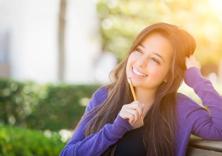 Attractive pensive mixed race female student with pencil sitting on campus bench