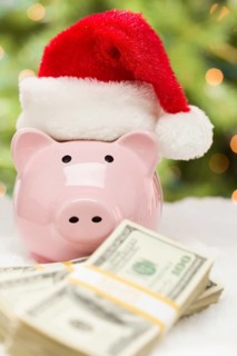Pink piggy bank wearing santa hat near stacks of hundreds of dollars of money on snowflakes
