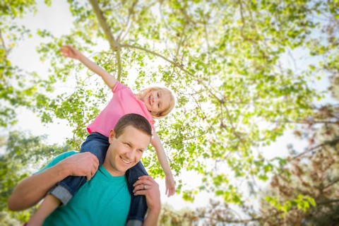 Cute young girl rides piggyback on her dads shoulders outside at the park