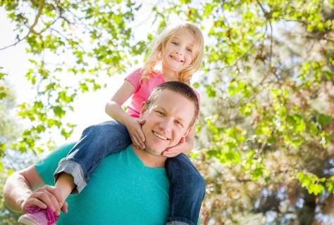 Cute young girl rides piggyback on her dads shoulders outside at the park