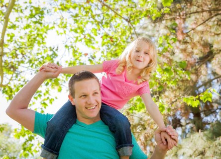 Cute young girl rides piggyback on her dads shoulders outside at the park