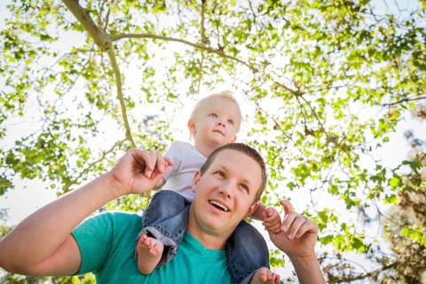 Cute young boy rides piggyback on his dads shoulders outside at the park