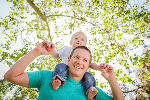 Cute young boy rides piggyback on his dads shoulders outside at the park