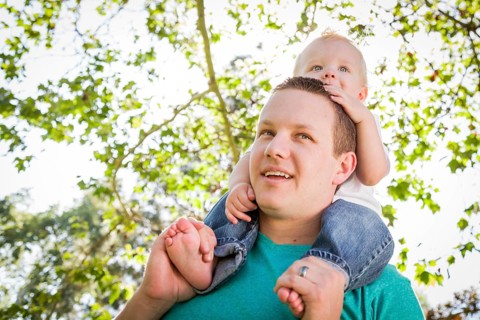 Cute young boy rides piggyback on his dads shoulders outside at the park