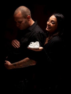 Fun mixed-race couple holding new white baby shoes against a black background under dramatic lighting
