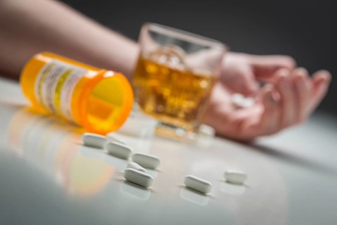 Hand laying on ground behind scattered prescription drugs and glass of alcohol