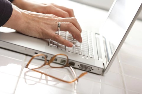 Female hands typing on the keyboard of the laptop with eyeglasses nearby