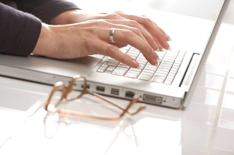 Female hands typing on the keyboard of the laptop with eyeglasses nearby