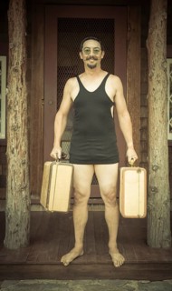 Goofy gentleman dressed in 1920's era swimsuit holding suitcases on porch of cabin