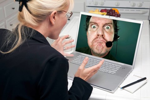 Shocked woman in kitchen using laptop with grumpy customer support man on screen