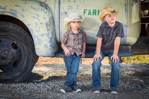 Two young boys wearing cowboy hats leaning against an antique truck in a rustic country setting