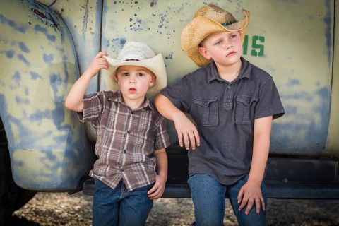 Two young boys wearing cowboy hats leaning against an antique truck in a rustic country setting