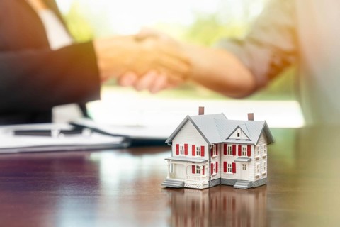 Real estate agent and customer sign contract papers and shake hands with small model home in front