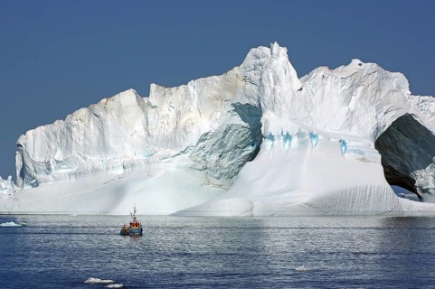 Small fishing boat in front of huge iceberg with two holes