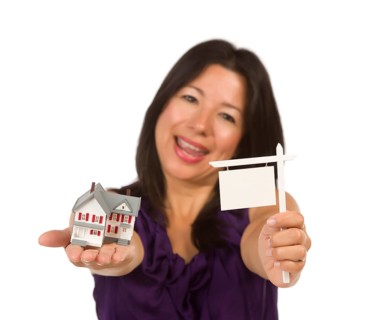 Multiethnic woman holding small blank real estate sign and house in hand isolated on white background