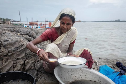 A woman sieves river water from wash bowls on a muddy embankment