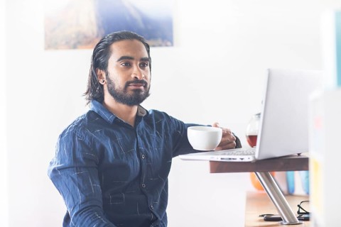 Young man with ponytail and checkered shirt drinking tea at home and working