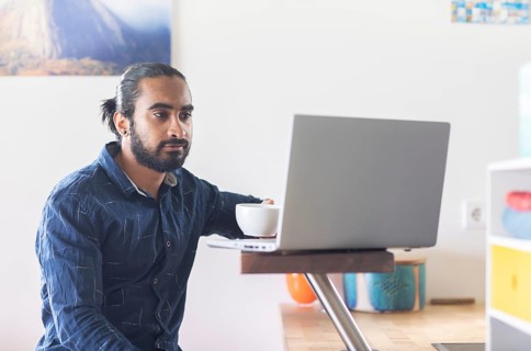 Young man with ponytail and checkered shirt drinking tea at home and working