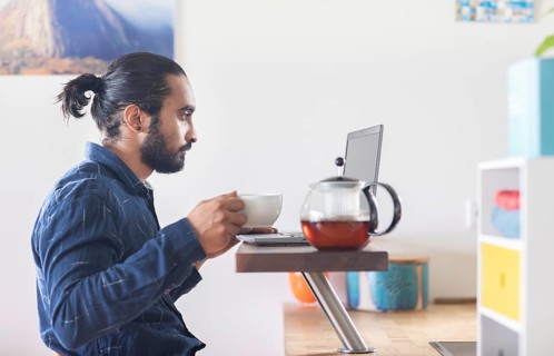 Young man with ponytail and checkered shirt drinking tea at home and working