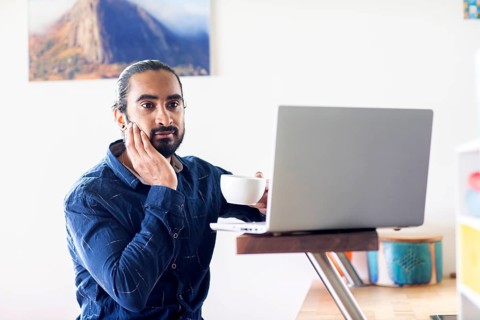 Young man with ponytail and checkered shirt drinking tea at home and working