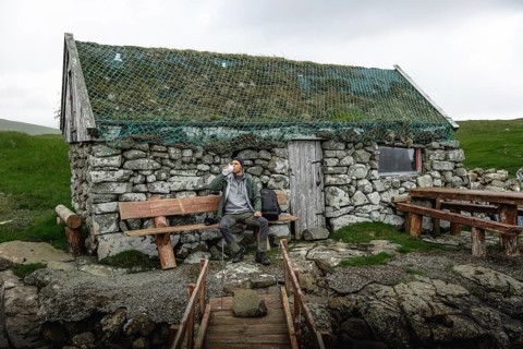 Hiker drinking water on a bench in front of a stone hut