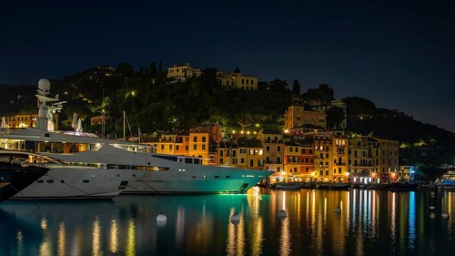 Luxury yachts and boats anchored in Portofino harbour at night in front of illuminated pastel-coloured house facades