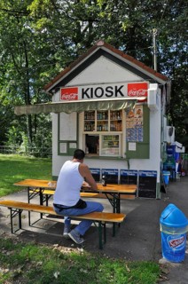 Man in white vest and jeans sits on bench in front of kiosk