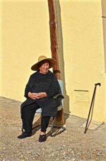 Old woman with straw hat and walking stick sitting in front of yellow house wall