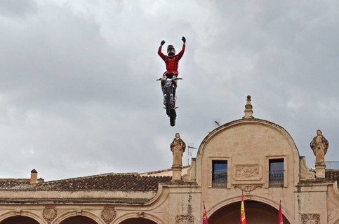 Motorbike performer flies through the air with arms raised above the roof of City Hall