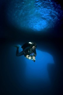 Diver dives with underwater lamp through rock grotto with blue light Blue Dome in Blue Hole of Gozo