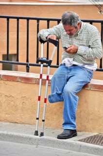 One-legged man with crutch sits on wall and looks into his mobile phone